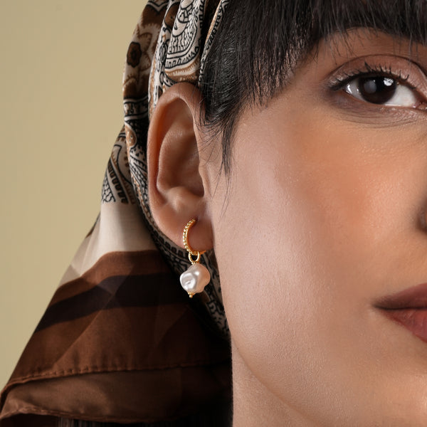 A close-up of a model wearing an elegant gold baroque pearl drop earring. The beaded huggie hoop is shown with her stylish brown paisley headscarf.