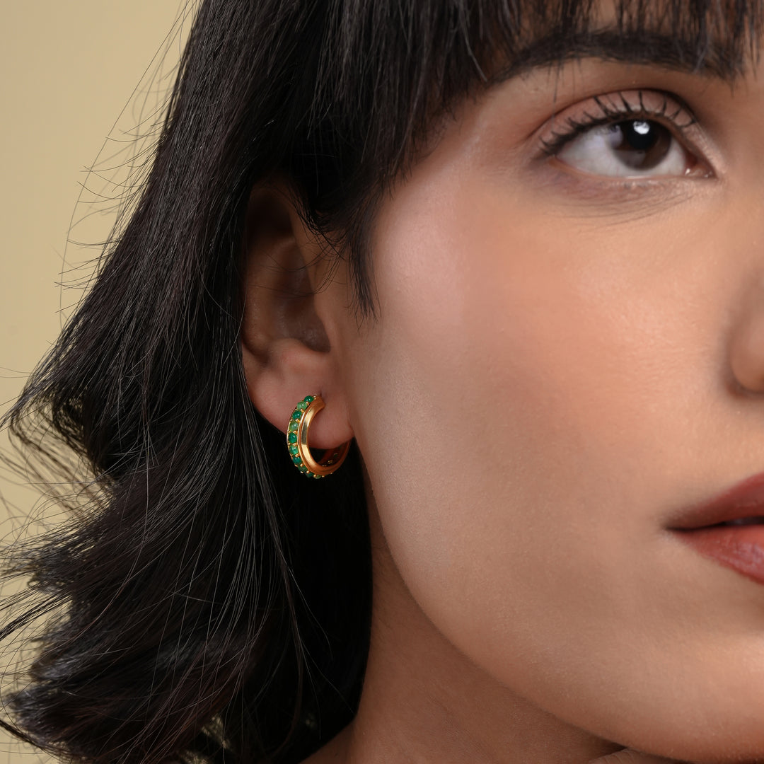 A close-up of a model wearing an elegant gold C-hoop earring, featuring a channel of sparkling emerald-green gemstones that hug her earlobe.