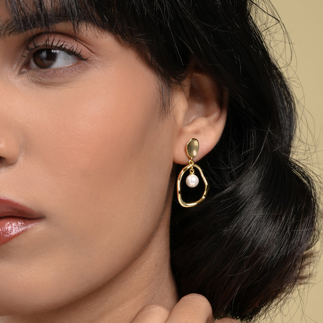 A close-up of a model showcasing a modern gold and pearl dangle earring. The unique design features a single pearl inside an organic, irregular-shaped gold hoop, highlighting its elegant drop and making it a perfect statement accessory.