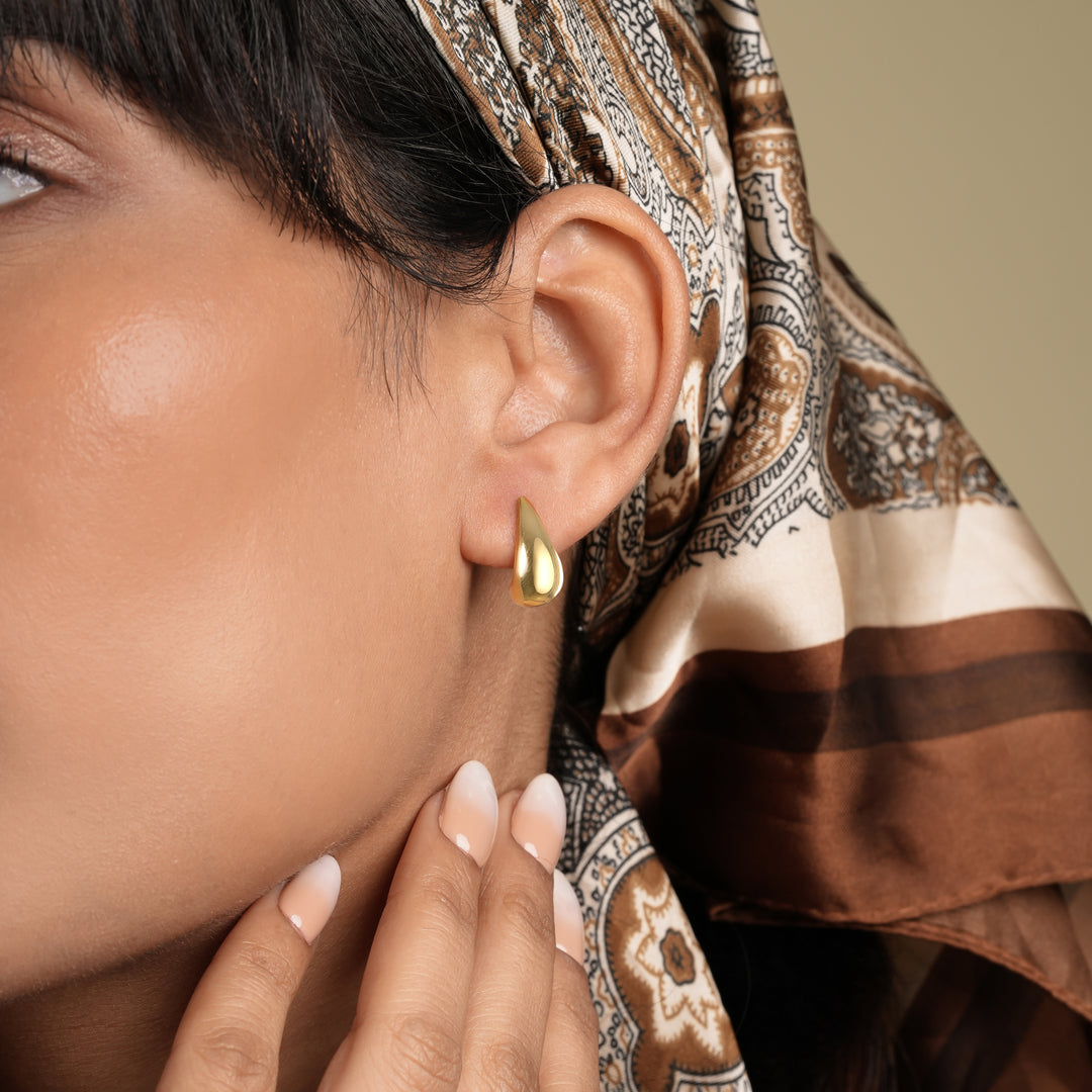 A close-up of a model wearing a minimalist gold teardrop stud earring from Zewory Jewellery. The elegant, polished droplet shape curves beautifully against her earlobe, showcased with a stylish paisley headscarf.