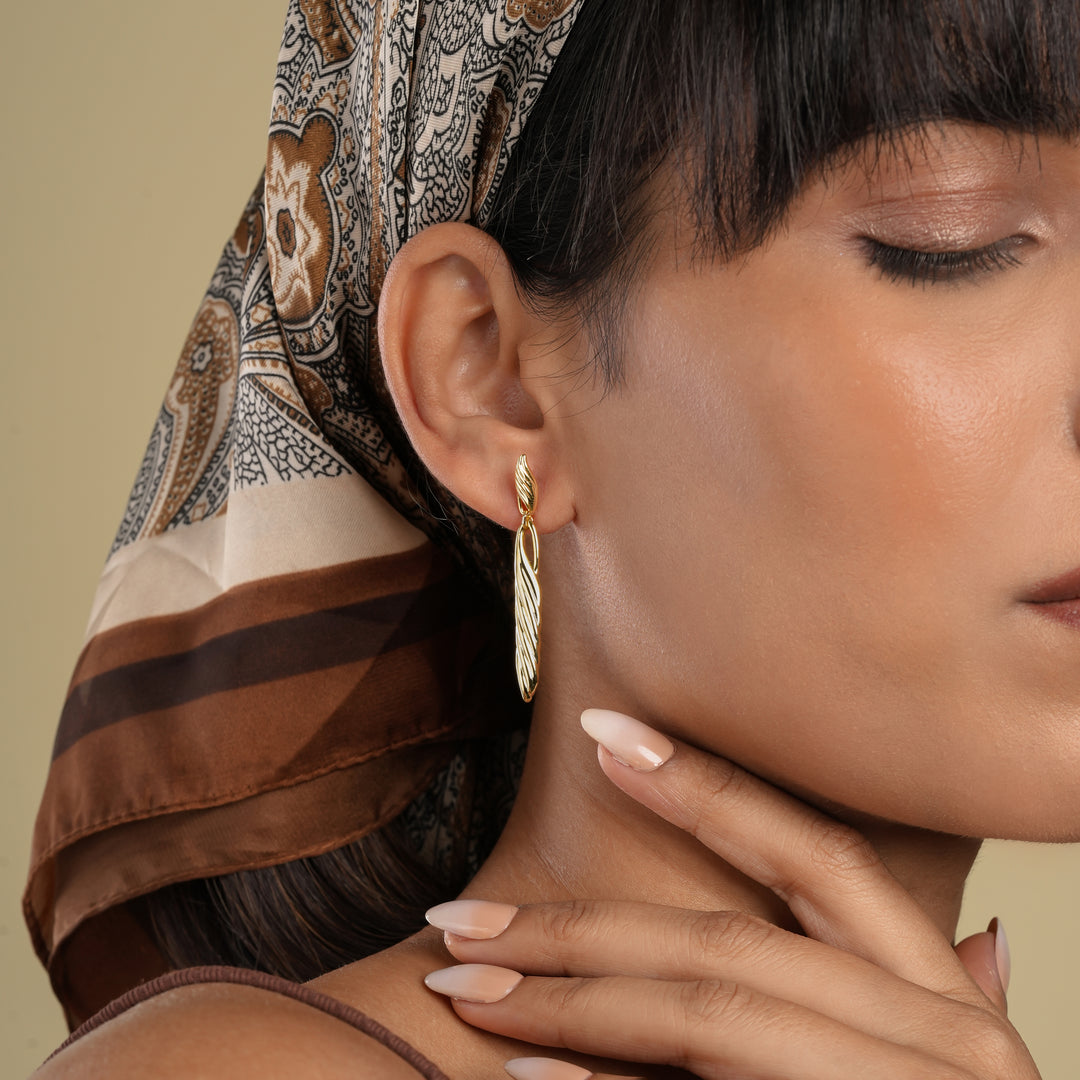 A close-up profile of a model wearing elegant, long twisted gold drop earrings. Her eyes are closed, and she is wearing a stylish brown and cream paisley headscarf, showcasing the high-fashion appeal of the statement earrings.