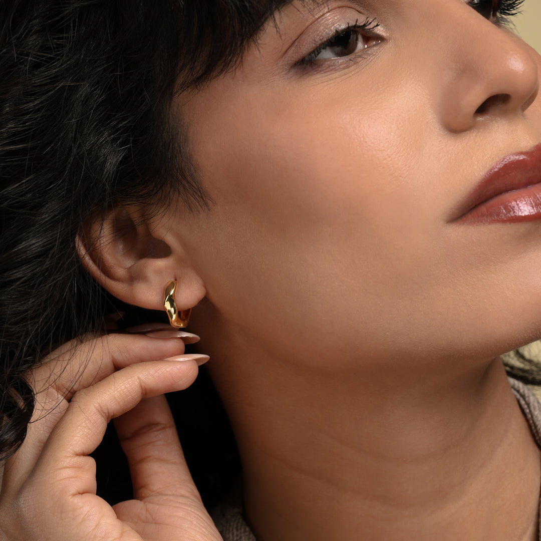 A close-up of a model wearing a modern, wavy gold huggie hoop earring from Zewory Jewellery. The small, sculptural hoop elegantly hugs her earlobe, showcasing a chic and minimalist accessory perfect for everyday style.