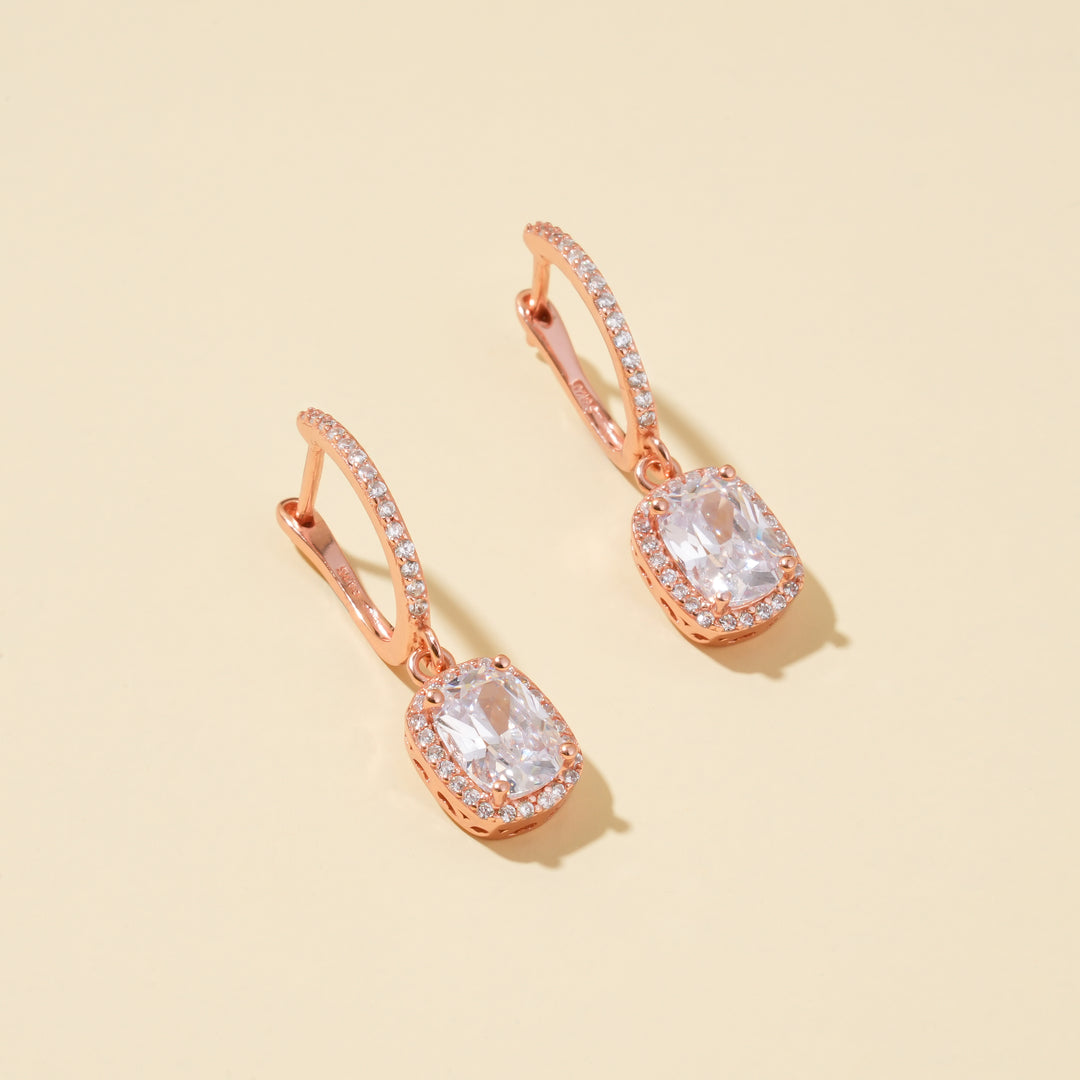 A clean product shot of a pair of rose gold halo drop earrings. The image highlights the sparkling, cushion-cut center stone and pavé crystal details on a neutral cream background.