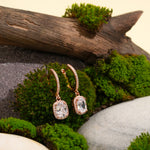 Load image into Gallery viewer, A pair of sparkling rose gold halo drop earrings from Zewory Jewellery. The elegant, cushion-cut crystal earrings are displayed in a natural setting of green moss, stone, and wood.
