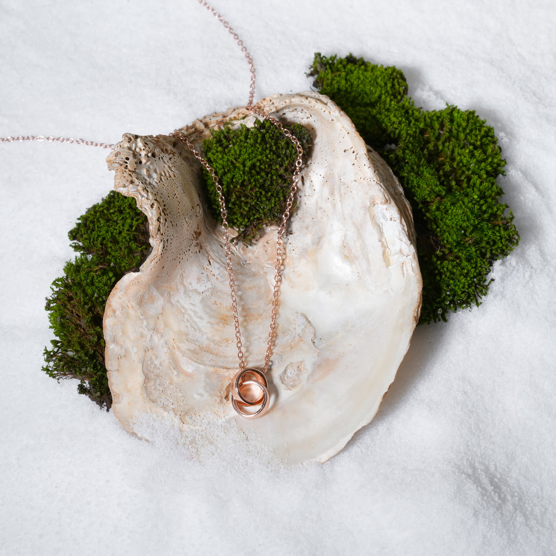 A beautiful rose gold necklace with an interlocking double-circle pendant, symbolizing connection and eternity. This stunning piece from Zewory Jewellery is artistically displayed in a large seashell on a bed of white sand and moss.