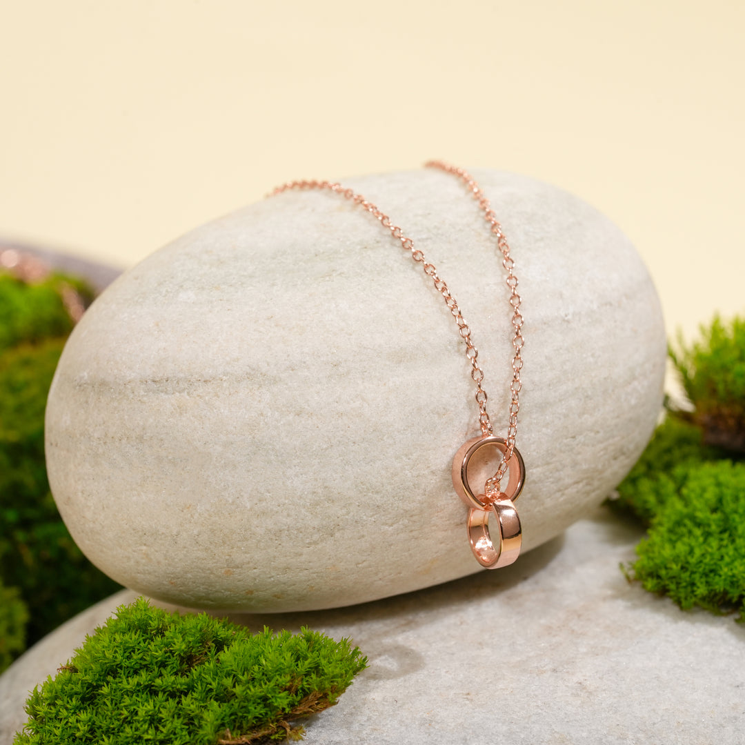 An elegant rose gold necklace from Zewory Jewellery, featuring a delicate double interlocking rings pendant. This minimalist piece is displayed on a smooth, light-grey stone surrounded by vibrant green moss for a natural and serene product shot.