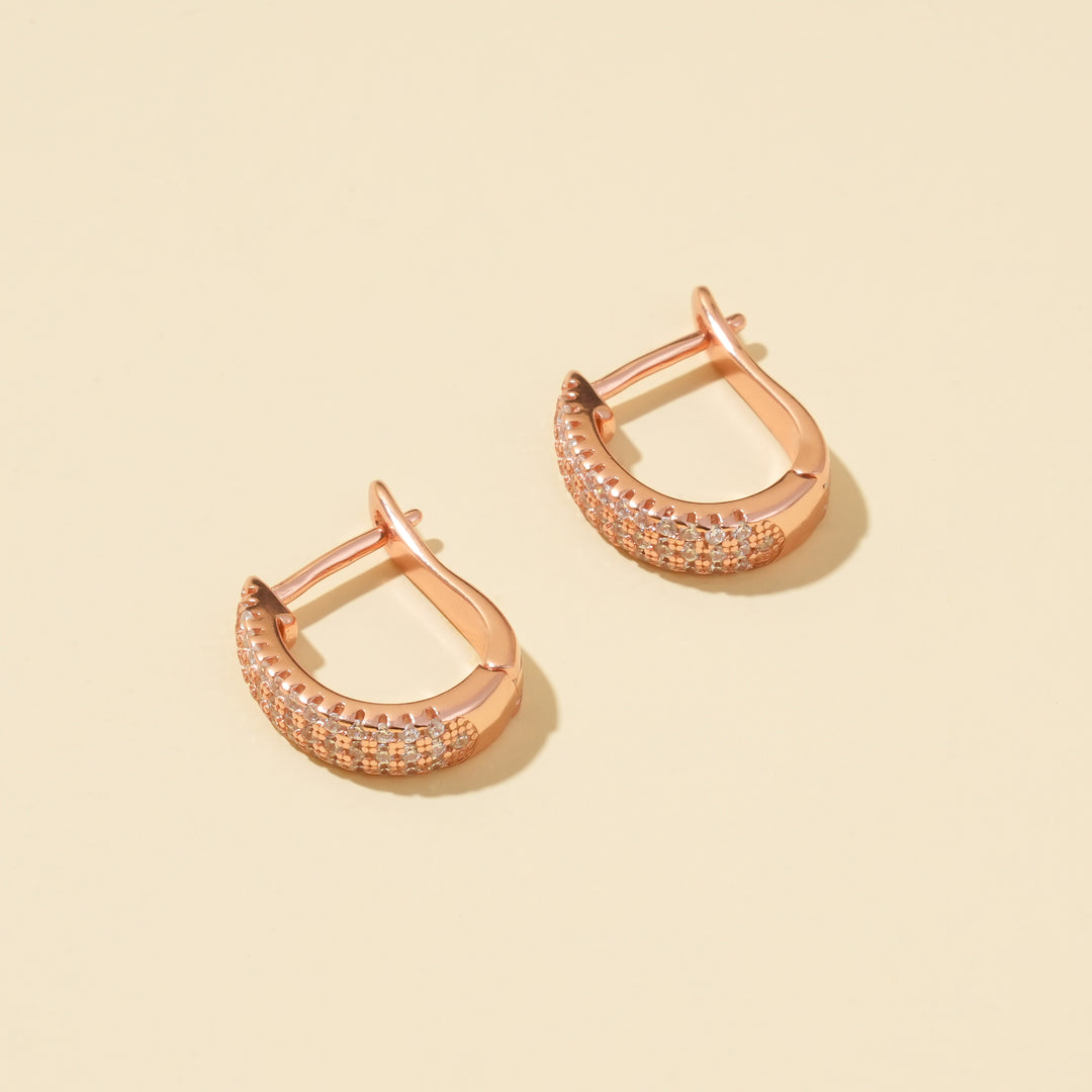A clean product shot of a pair of elegant rose gold hoop earrings on a neutral cream background. The image clearly displays the multiple rows of sparkling pavé-set crystals that cover the front of the small, U-shaped huggie hoops.