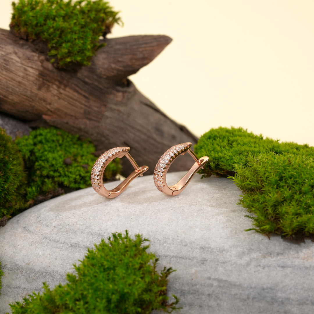 A pair of brilliant rose gold pavé hoop earrings from Zewory Jewellery. The sparkling, crystal-covered design is showcased in a natural setting on a grey stone, surrounded by vibrant green moss and weathered wood.