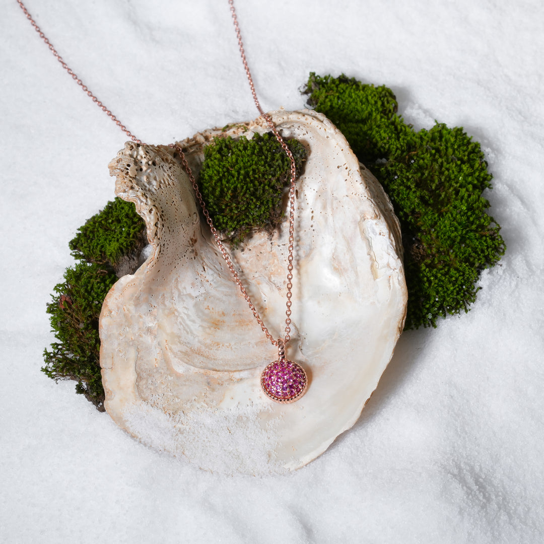 A luxury product shot of a rose gold necklace featuring a brilliant, round pendant covered in pink pavé-set stones. The necklace is artfully displayed inside a large seashell on a bed of white sand and moss.