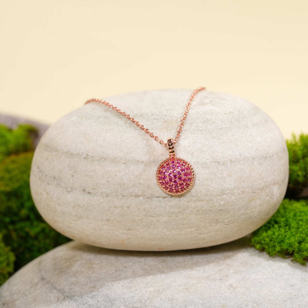 An elegant rose gold necklace with a vibrant pink pavé disc pendant from Zewory Jewellery. The piece is beautifully displayed on a smooth, light-coloured stone, surrounded by fresh green moss for a natural aesthetic.