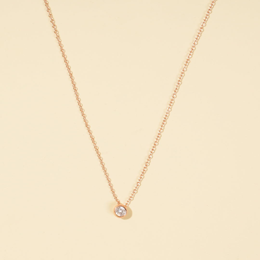 A clean, minimalist product shot of a rose gold solitaire necklace on a neutral cream background. The image clearly displays the delicate chain and the classic, round brilliant-cut stone in a modern bezel setting.