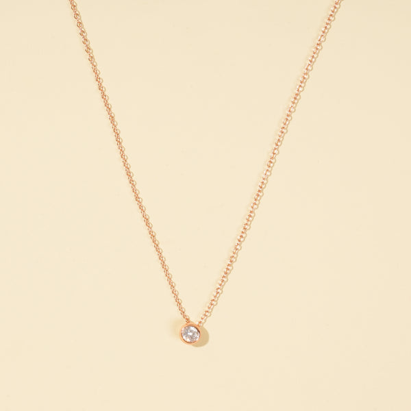 A clean, minimalist product shot of a rose gold solitaire necklace on a neutral cream background. The image clearly displays the delicate chain and the classic, round brilliant-cut stone in a modern bezel setting.