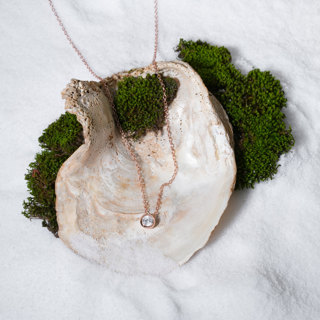 A luxurious product shot of a rose gold solitaire necklace from Zewory Jewellery. This beautiful and timeless piece, featuring a sparkling bezel-set stone, is artistically arranged inside a large seashell on a bed of white sand and moss.
