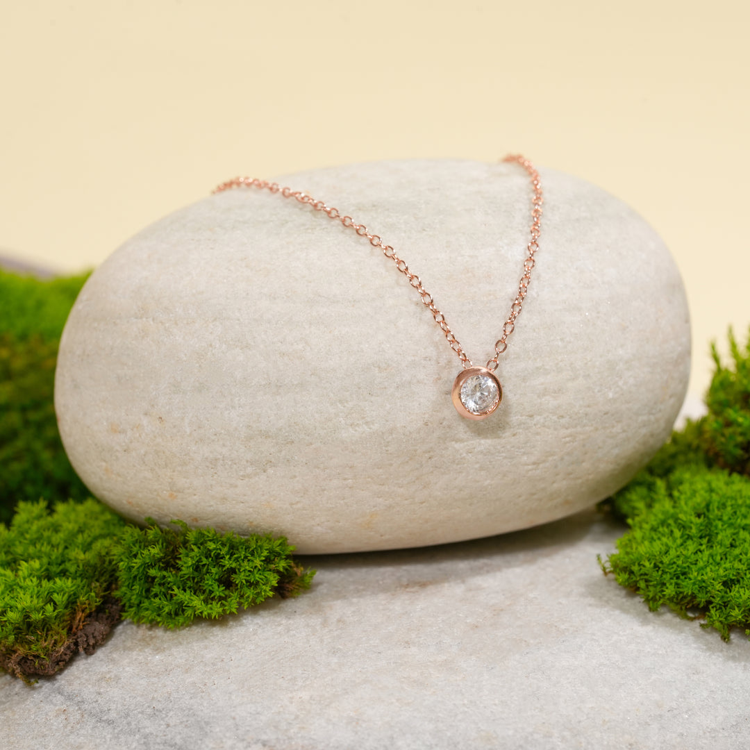 A delicate rose gold solitaire necklace from Zewory Jewellery, displayed in a serene natural setting. The minimalist, bezel-set crystal pendant is draped over a smooth, light-grey stone and surrounded by fresh green moss.