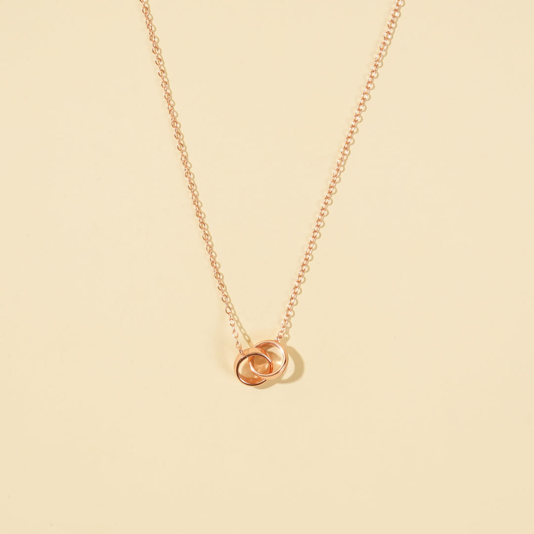 An elegant rose gold necklace from Zewory Jewellery, featuring a delicate double interlocking rings pendant. This minimalist piece is displayed on a smooth beige plain background.
