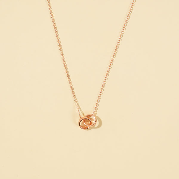 An elegant rose gold necklace from Zewory Jewellery, featuring a delicate double interlocking rings pendant. This minimalist piece is displayed on a smooth beige plain background.
