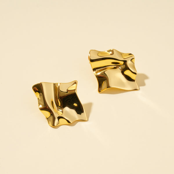 A clean product shot of a pair of abstract, sculptural gold stud earrings. The image highlights the modern, crumpled-metal design and high-polish finish of the statement earrings on a neutral cream background.