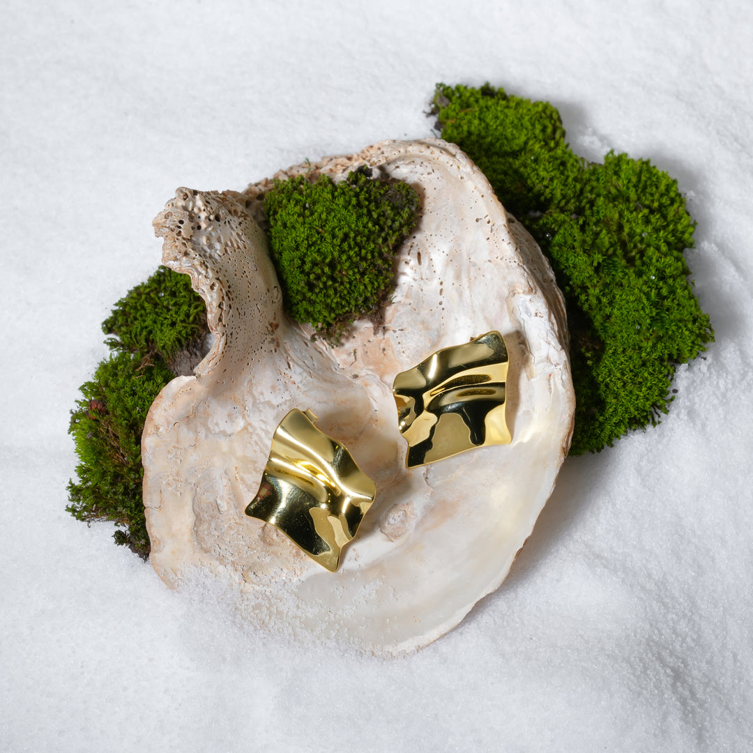 A luxurious product shot of sculptural gold statement stud earrings from Zewory Jewellery. The pair of modern, abstract earrings with a molten-metal look are artistically displayed inside a large seashell on a bed of white sand and moss.