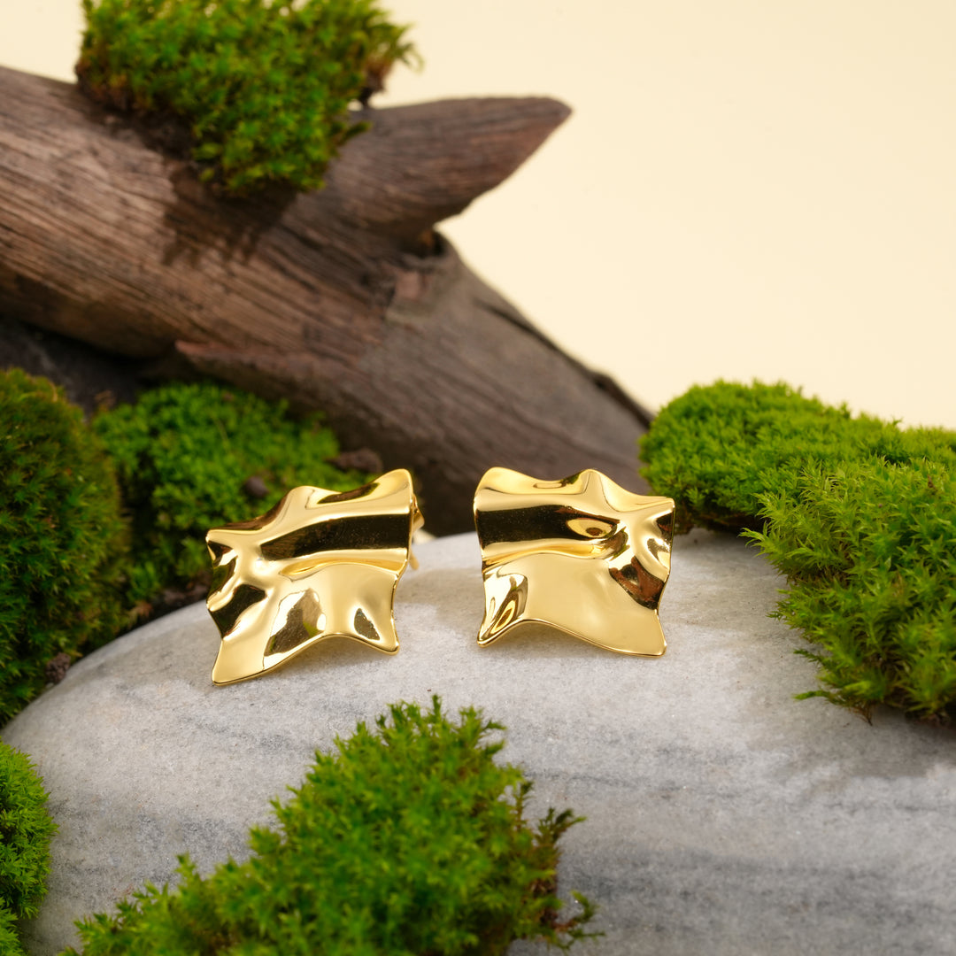 A pair of modern, wavy gold statement stud earrings from Zewory Jewellery. The artistic, sculptural design is showcased in a natural setting on a grey stone, surrounded by vibrant green moss and weathered wood.