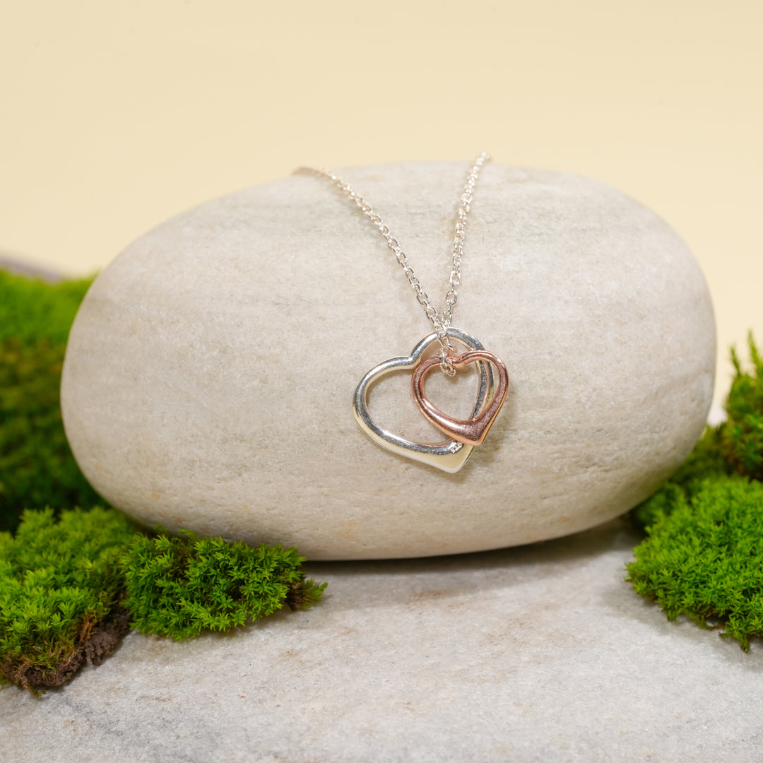 An elegant two-tone double heart necklace from Zewory Jewellery, displayed in a serene natural setting. The minimalist silver and rose gold interlocking heart pendant is draped over a smooth, light-grey stone and surrounded by fresh green moss.