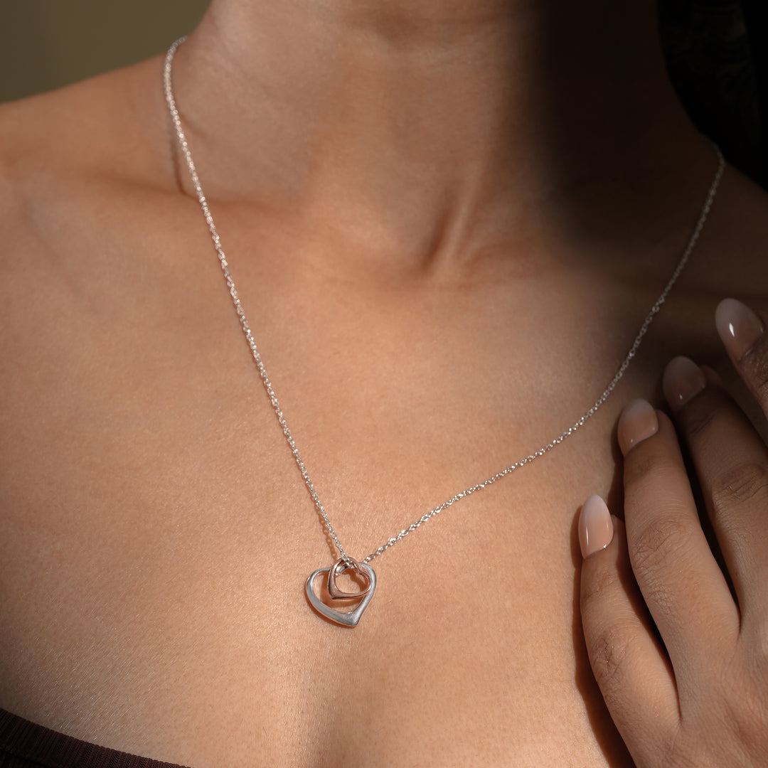A close-up of a model wearing an elegant two-tone interlocking hearts necklace from Zewory Jewellery. The romantic pendant, featuring a large silver open heart and a smaller rose gold open heart, rests gracefully against her skin.