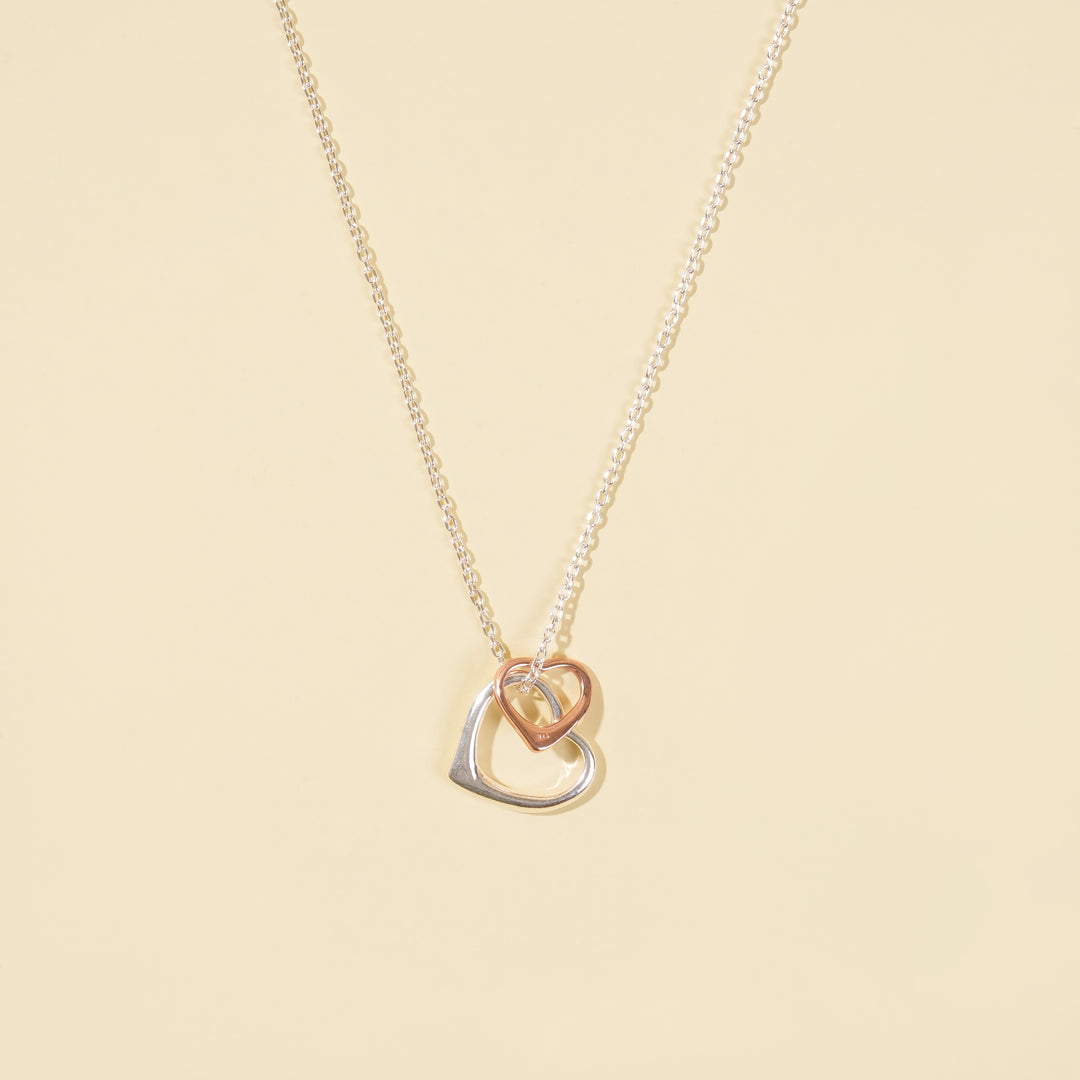 A clean, minimalist product shot of a two-tone interlocking hearts necklace on a neutral cream background. The image clearly displays the delicate silver chain with its mixed-metal, double-heart pendant.