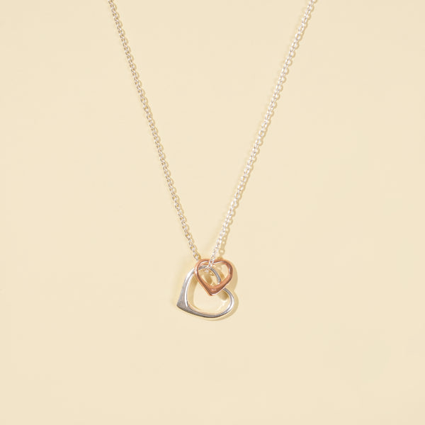 A clean, minimalist product shot of a two-tone interlocking hearts necklace on a neutral cream background. The image clearly displays the delicate silver chain with its mixed-metal, double-heart pendant.
