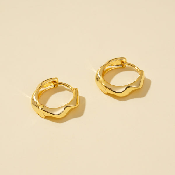 A clean product shot of a pair of minimalist gold huggie hoops with a modern, irregular wavy design. The image clearly displays the sculptural shape and high-polish finish of the earrings against a neutral cream background.