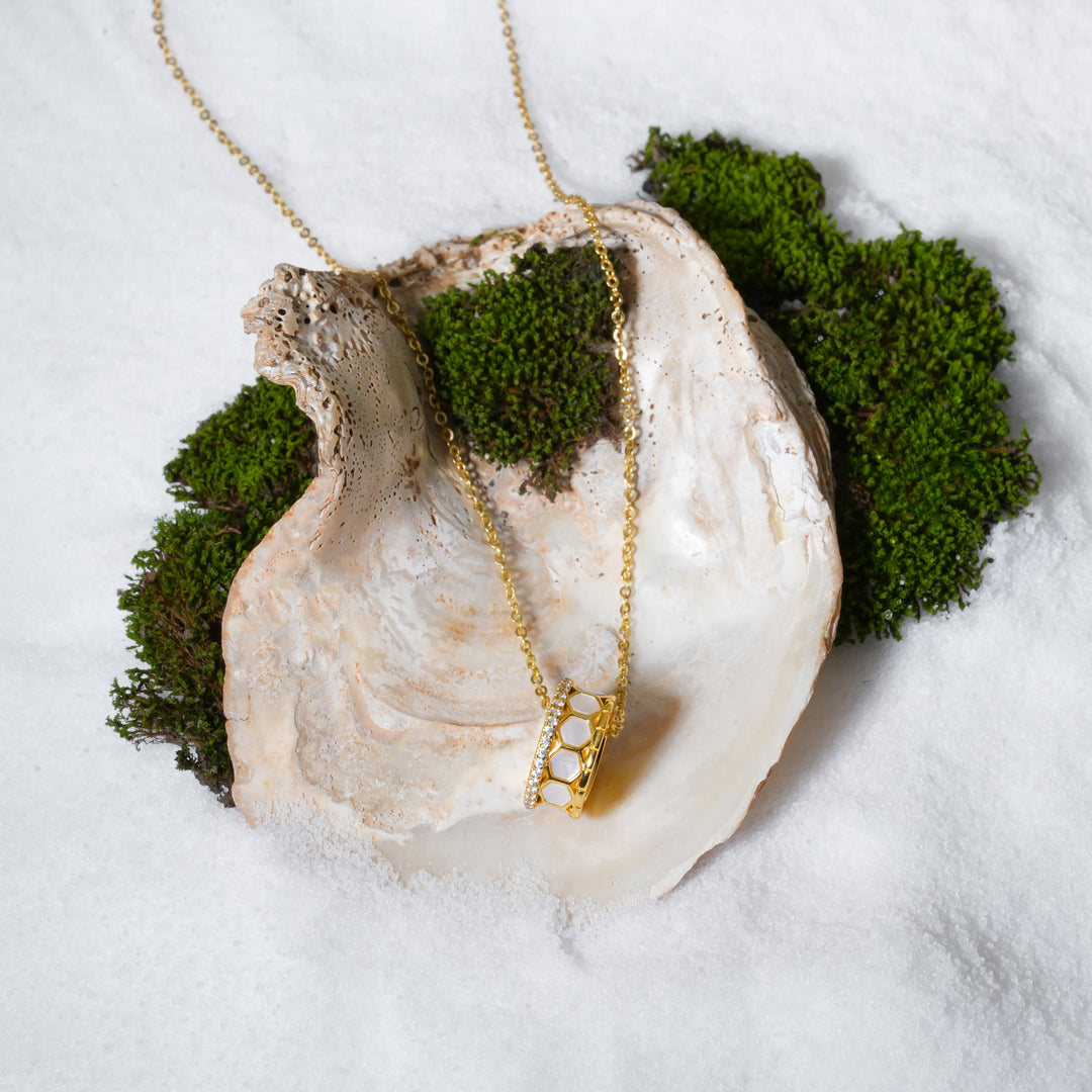 A luxurious product shot of a gold honeycomb barrel necklace from Zewory Jewellery. This chic pendant, with its mother-of-pearl inlays and pavé crystal accents, is artistically arranged inside a large seashell on a bed of white sand and moss.