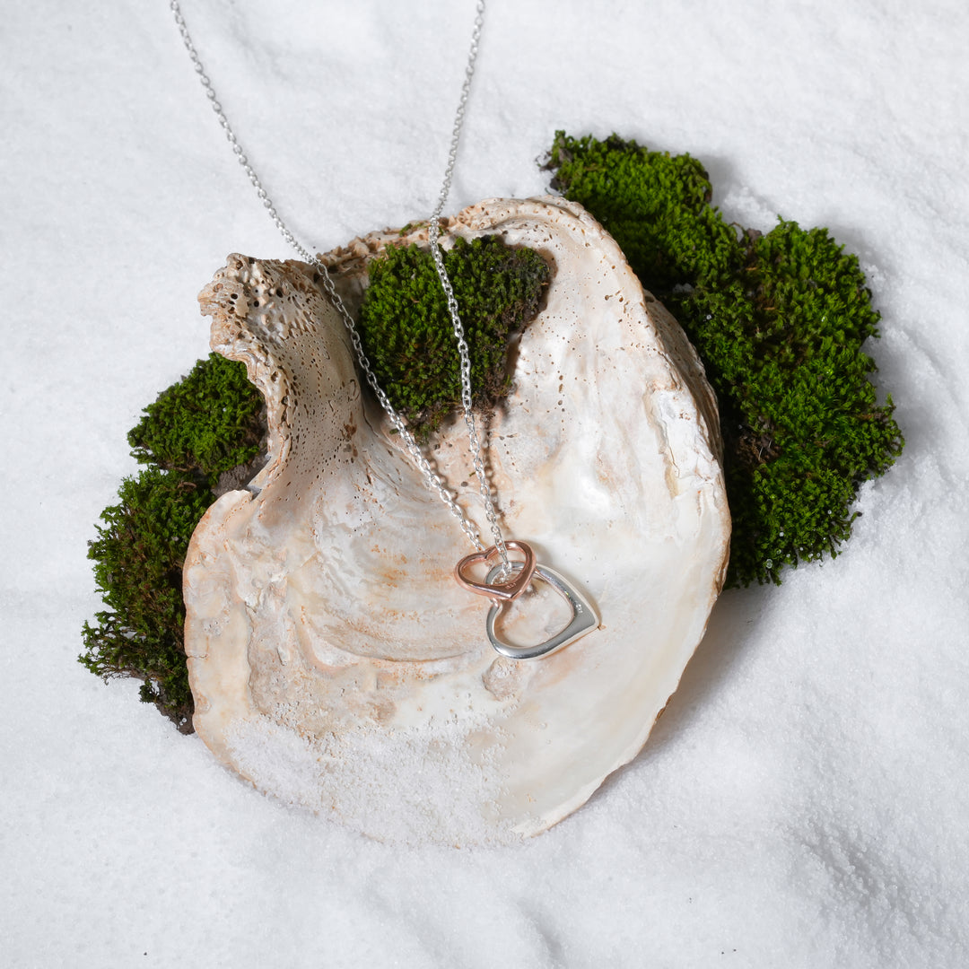 A luxurious product shot of a silver and rose gold interlocking hearts necklace. This beautiful and romantic piece of mixed-metal jewellery from Zewory is artistically arranged inside a large seashell on a bed of white sand and moss.